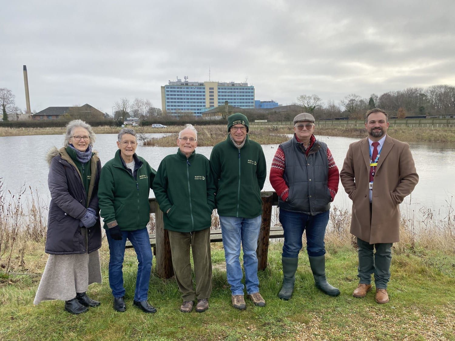 Trust volunteers and guests beside the lake at Dion's Wood with Pilgrim Hospital behind